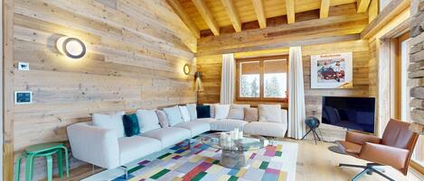 A cozy log cabin interior with a large white sectional sofa, a colorful rug, and a stone wall with a poster.