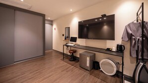 Ein modernes Hotelzimmer mit einem großen Flachbildfernseher, einem Schreibtisch mit Computer, einer kleinen Sitzecke, einem Föhn und einem Wäscheständer.