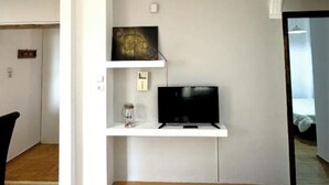 Et værelse med trægulv, et sort tv på en hvid hylde, en lille glasbeholder og en hvid væg med en døråbning.