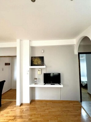A room with a wooden floor, a black TV on a white shelf, a small glass container, and a white wall with a door frame. - An Athenian gem by the Acropolis! (Athina)