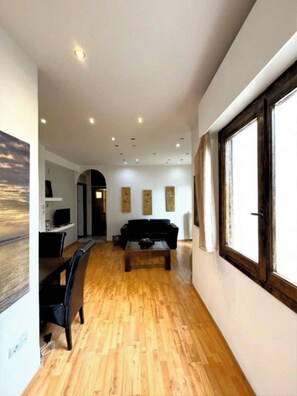 A spacious living area with a wooden floor, a black sofa, a coffee table, and a TV on a white stand. There are three paintings on the wall and a window on the right side. - An Athenian gem by the Acropolis! (Athina)
