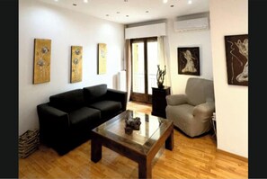 A living room with a black sofa, a grey armchair, and a coffee table. There are three paintings on the wall and a window with curtains. - An Athenian gem by the Acropolis! (Athina)