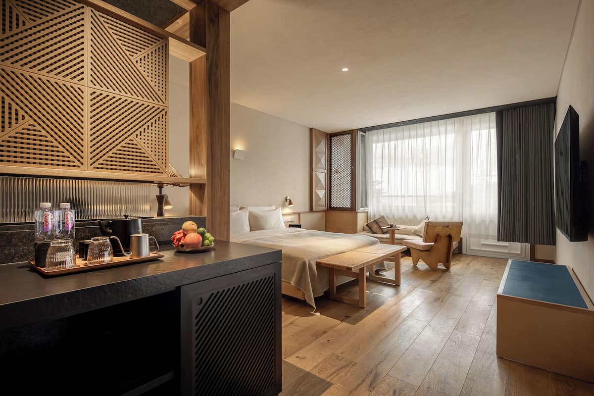 Una habitación de hotel moderna con piso de madera, una cama, un escritorio, un televisor y un ventanal grande con cortinas.