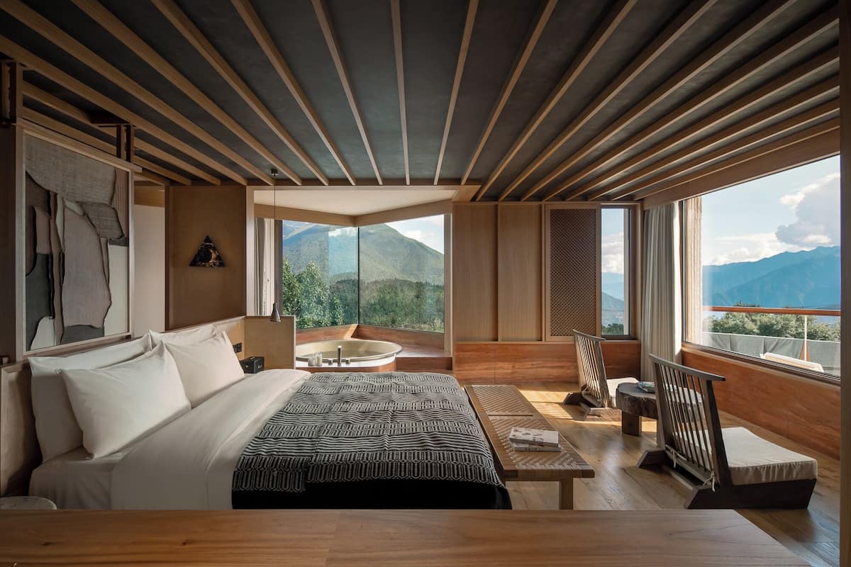 Un dormitorio moderno con una cama grande, un banco de madera, una mesita pequeña y vistas a montañas a través de amplios ventanales.