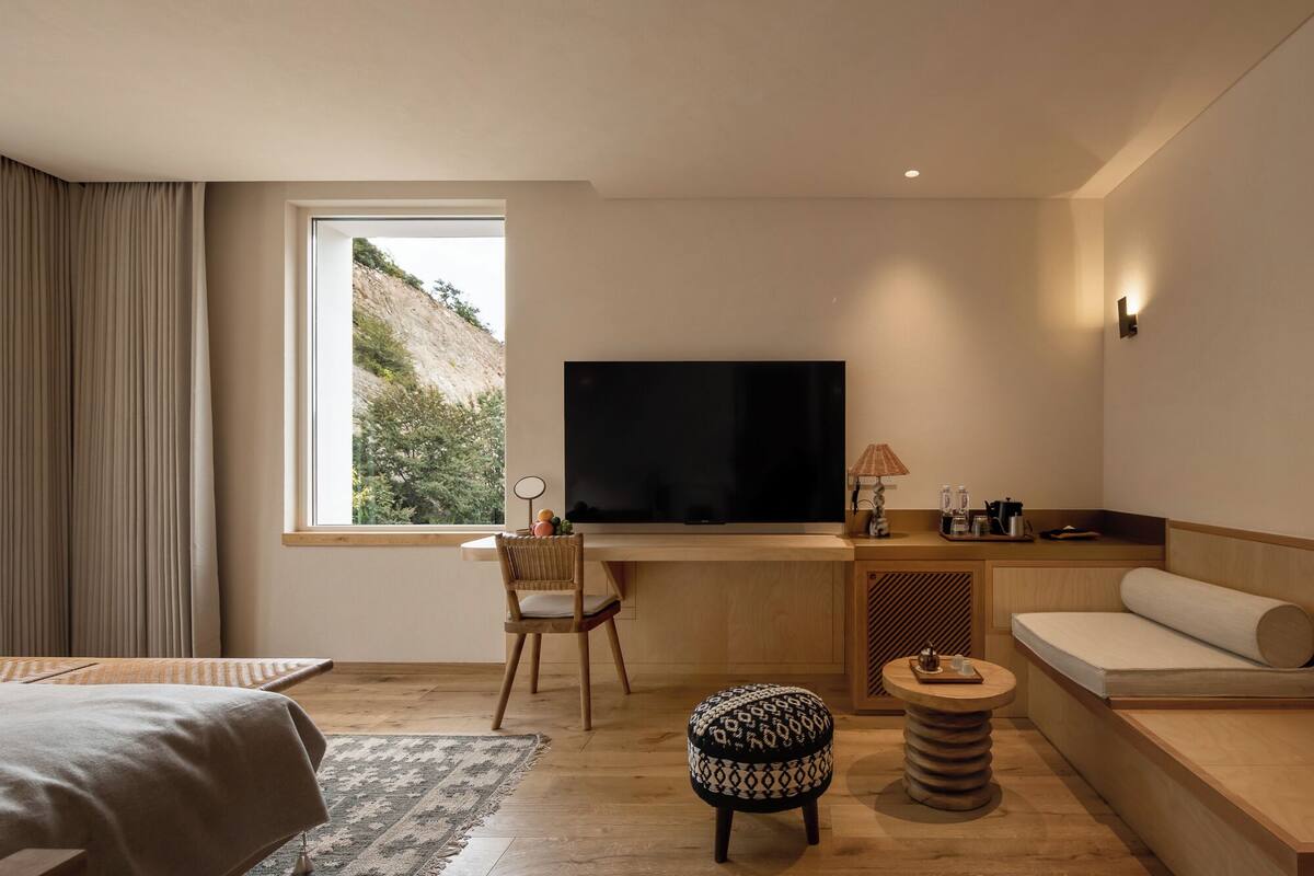 Habitación de hotel moderna con un ventanal grande, un televisor de pantalla plana, un sofá, un escritorio y una mesita redonda.