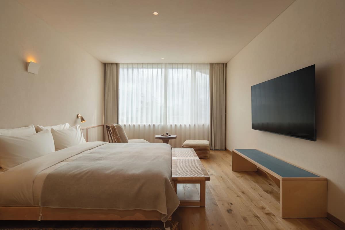 Habitación de hotel moderna con una cama grande, una mesita auxiliar pequeña, un banco y un televisor de pantalla plana montado en la pared.