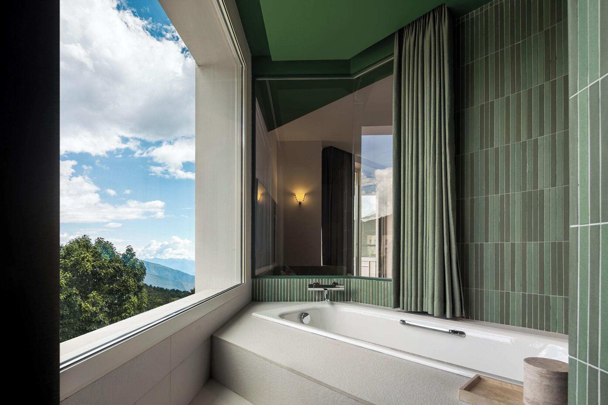 Un baño moderno con una bañera grande, una ventana con vistas a montañas y una pared con azulejos verdes.