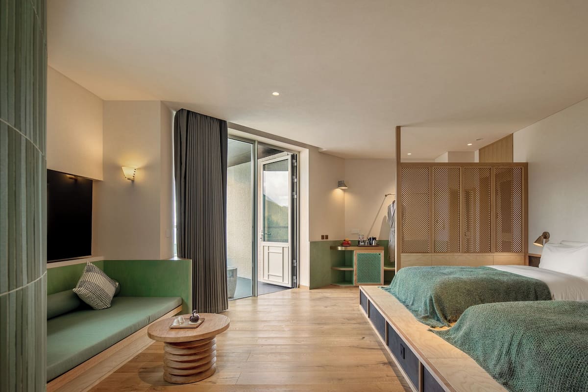 Una habitación de hotel moderna con un sofá verde, una mesa de centro de madera, una cama con manta verde, un separador de madera y vistas al exterior.