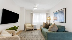 Un salon doté d’un téléviseur à écran plat, d’un ventilateur de plafond, d’un canapé vert, d’un fauteuil en osier, d’une petite table ronde et d’un grand tableau encadré accroché au mur.