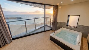 Un baño moderno con una bañera grande, un balcón con paredes de vidrio y vista al océano, y un espejo amplio.