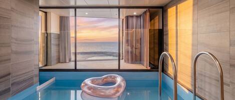 A modern indoor pool with a clear view of the ocean at sunset, a glass partition, and a metal ladder.