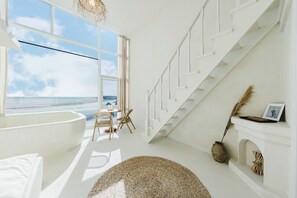 Amplia habitación con un gran ventanal con vistas al mar, una escalera blanca y una chimenea con decoración rústica.