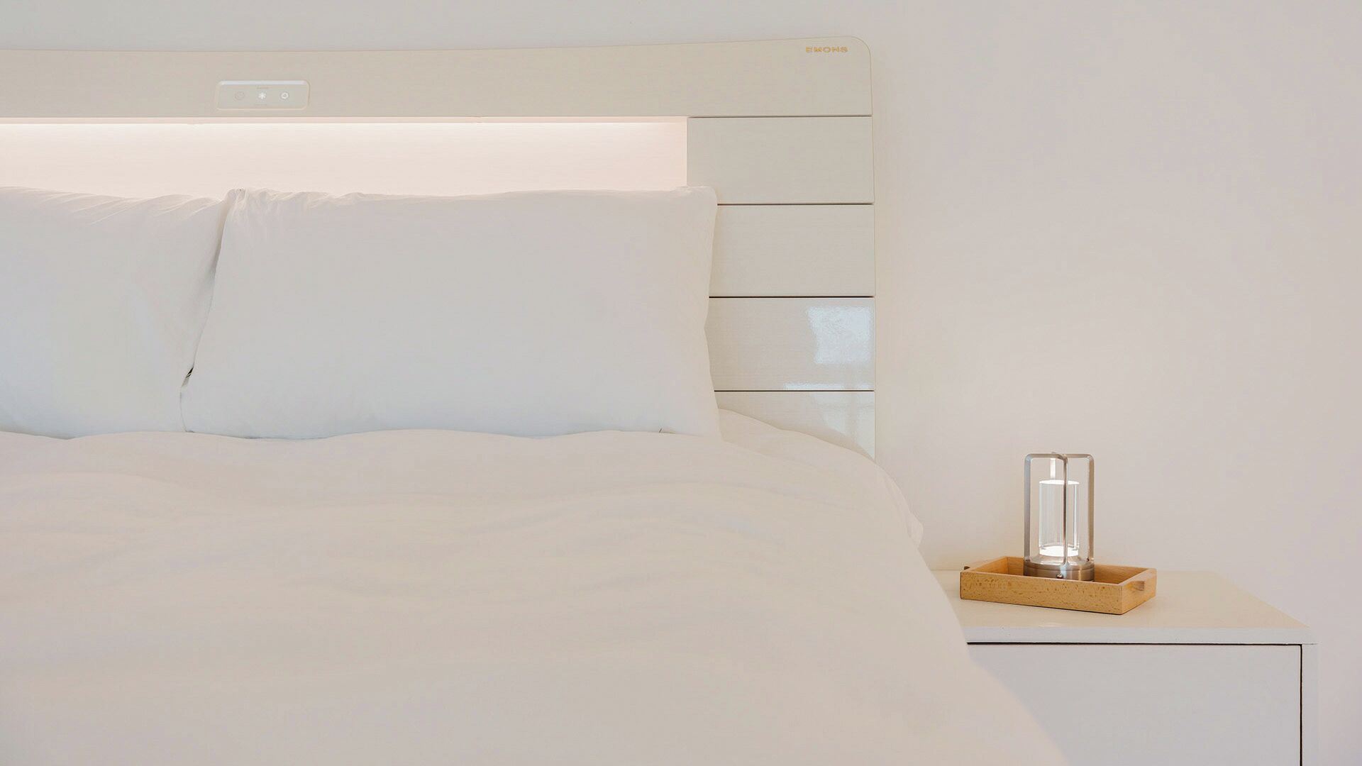 A bed with white bedding and pillows, a bedside table with a clear glass container, and a white headboard with a control panel.