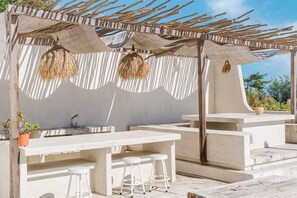 Un patio al aire libre con barra de concreto, taburetes blancos y una pérgola con cestas colgantes.
