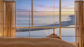 A room with a large window overlooking a beach, curtains, and a bed.