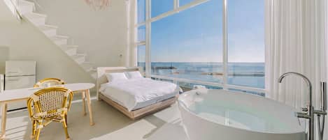 A modern bedroom with a large window, a bed, a sitting area with a table and chairs, and a bathtub.