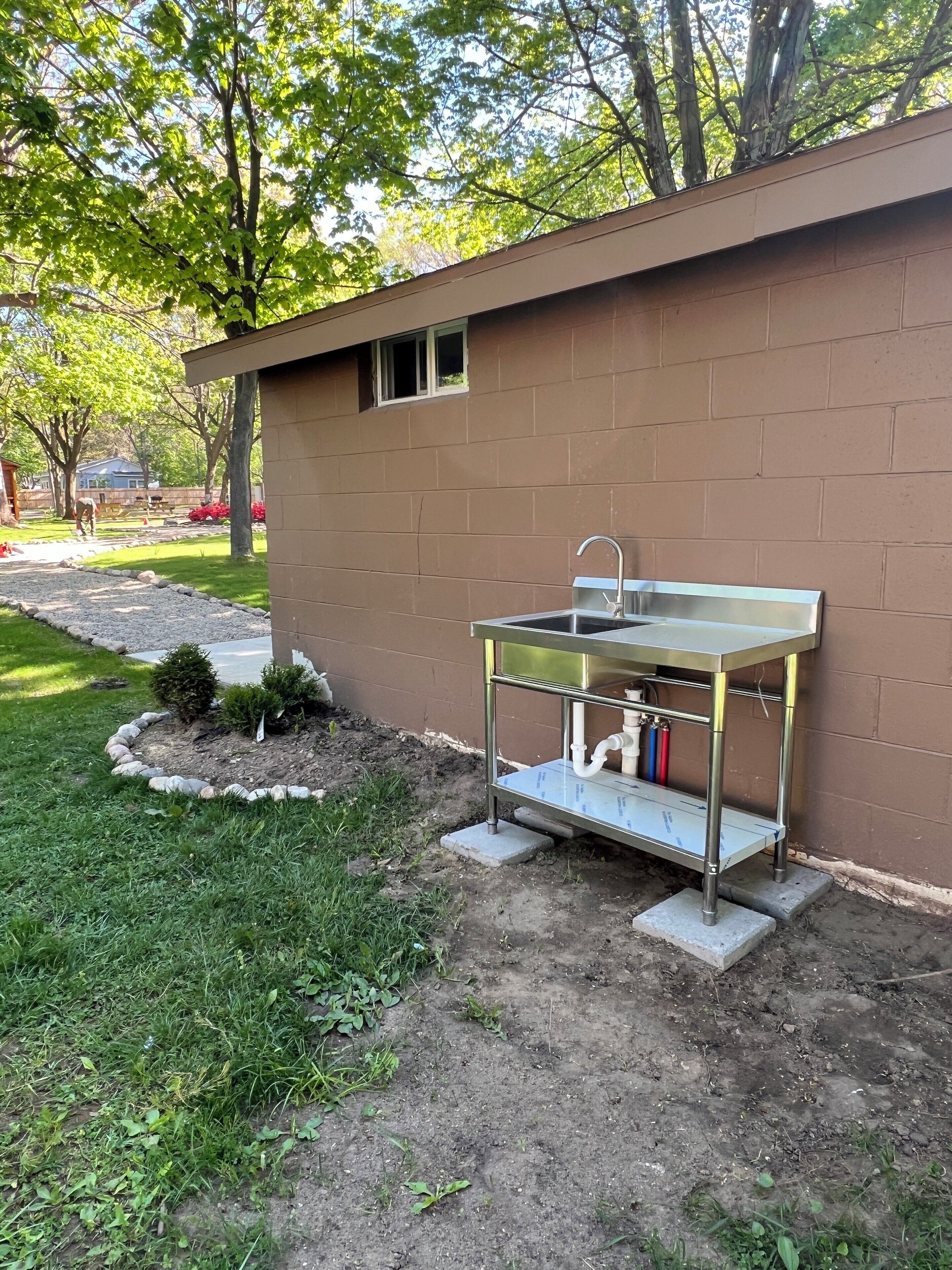 A small outdoor kitchen area with a sink and a window, surrounded by a brick wall and a grassy area with trees.