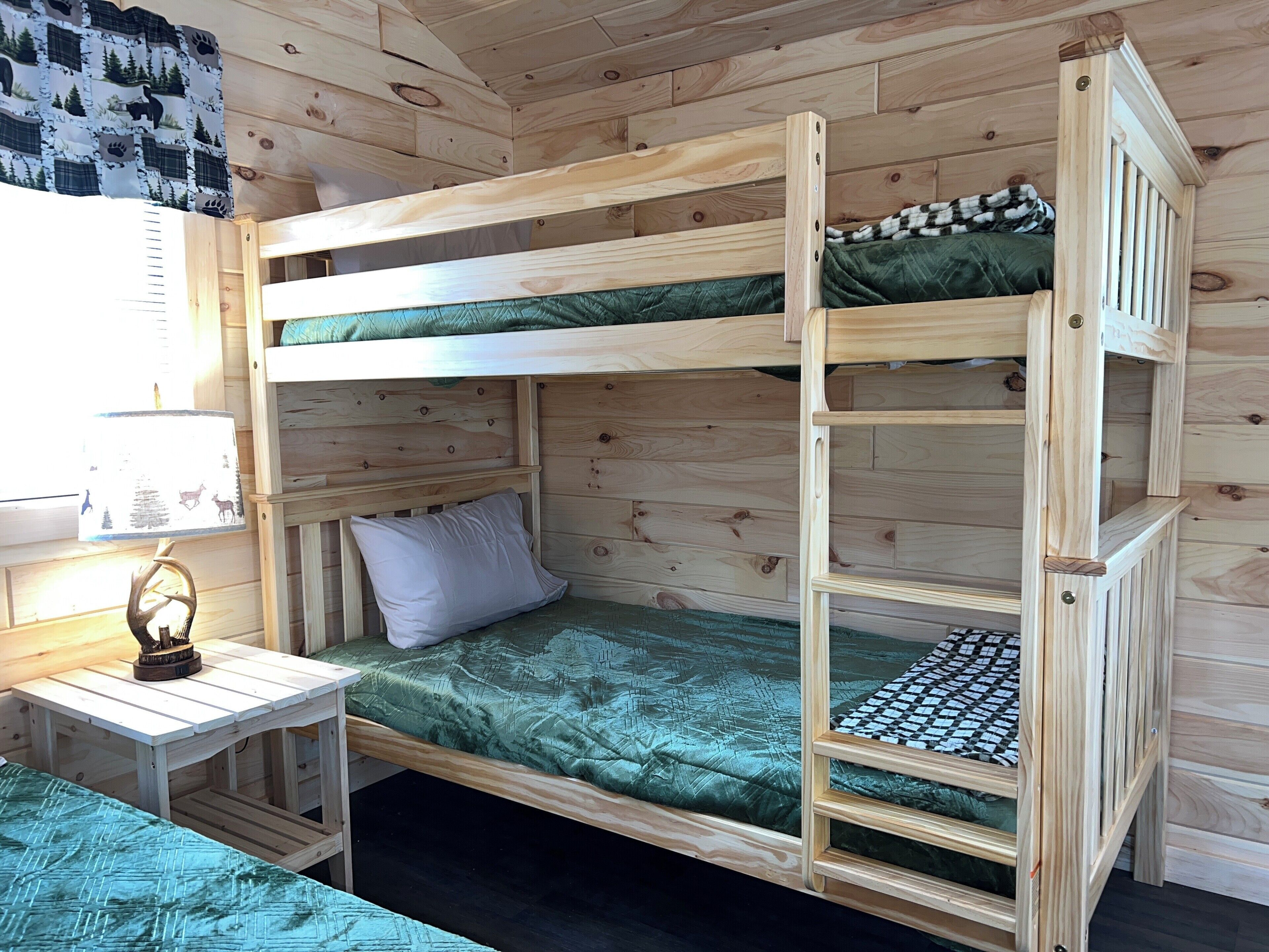 Habitación con literas, suelo de madera, una mesita con lámpara y una ventana con cortina.