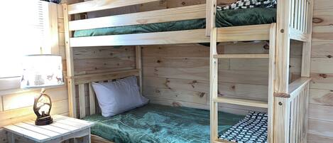 Habitación con literas, suelo de madera, una mesita con lámpara y una ventana con cortina.