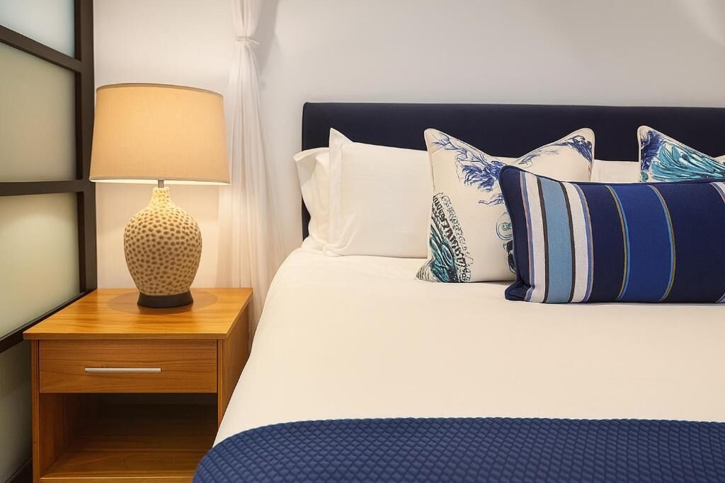 A neatly made bed with a blue quilt, a wooden nightstand with a lamp, and decorative pillows.