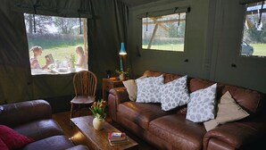 A cozy living room with a brown leather sofa, a wooden coffee table, and a chair. There are two windows with curtains, a lamp on the table, and a view of the outdoors.