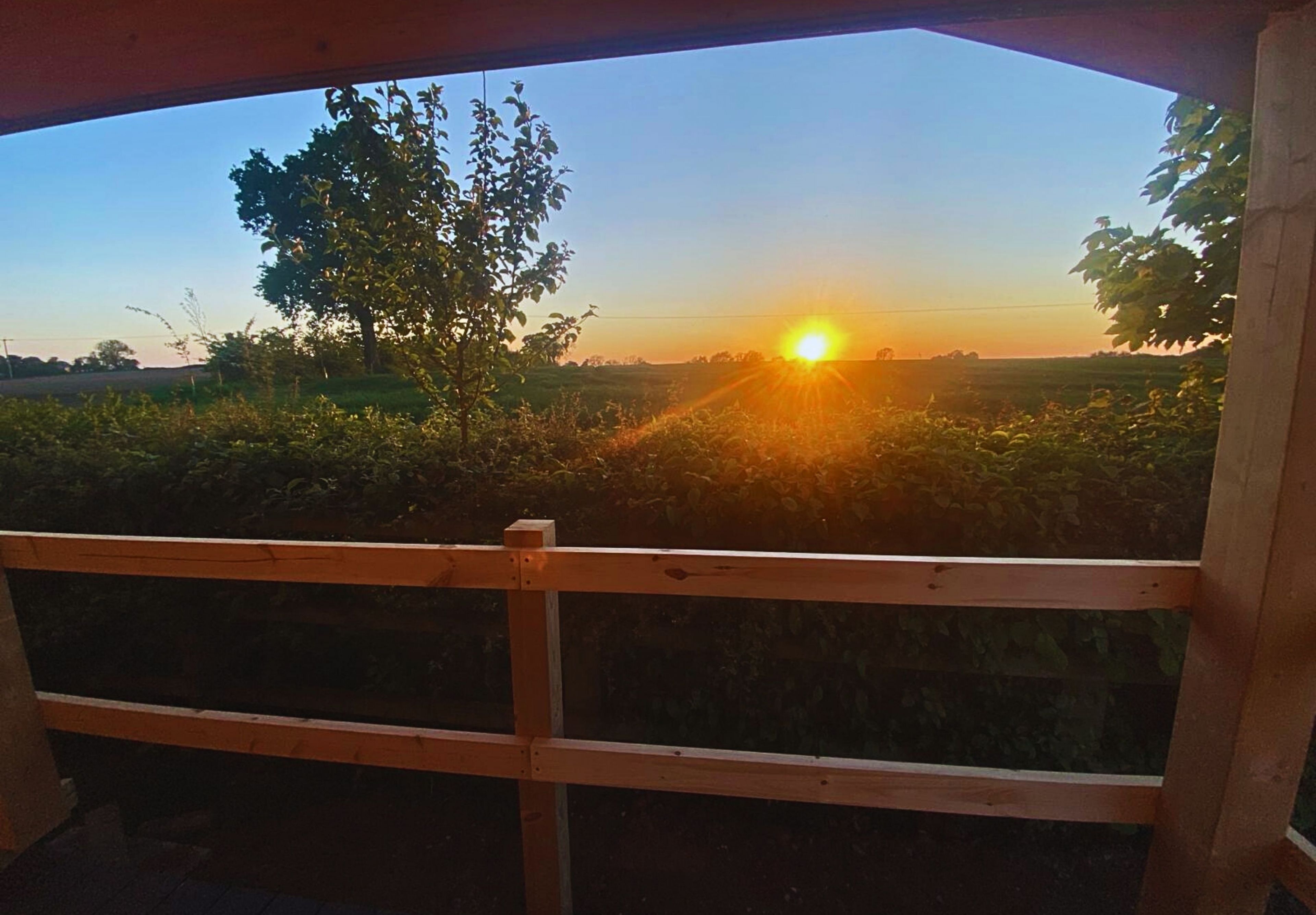 Une vue depuis une fenêtre à cadre en bois, donnant sur un champ baigné par les couleurs d’un coucher de soleil.