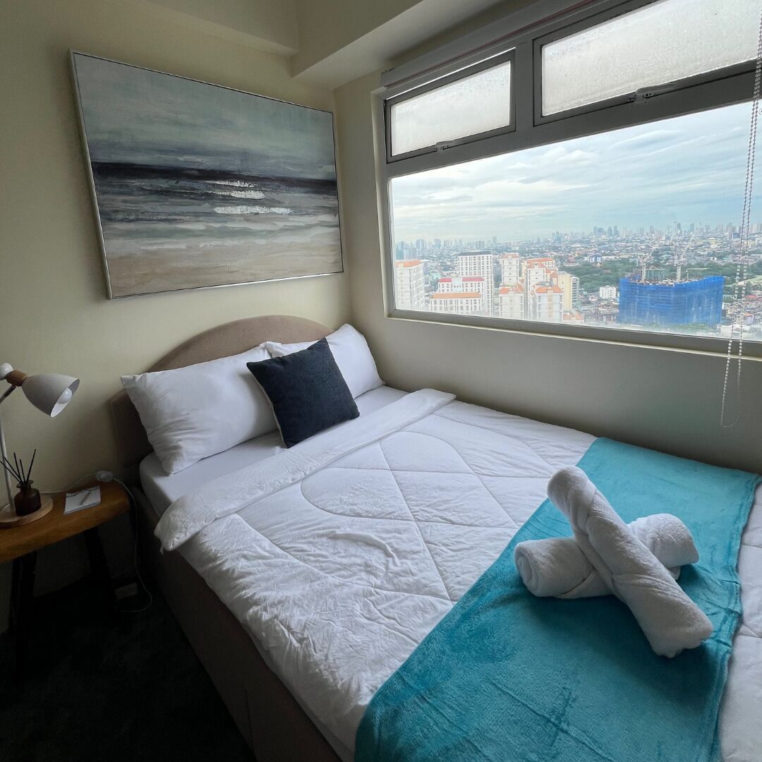 Une chambre d’hôtel avec un lit, une fenêtre donnant sur la ville, une lampe de chevet et un tableau au mur.