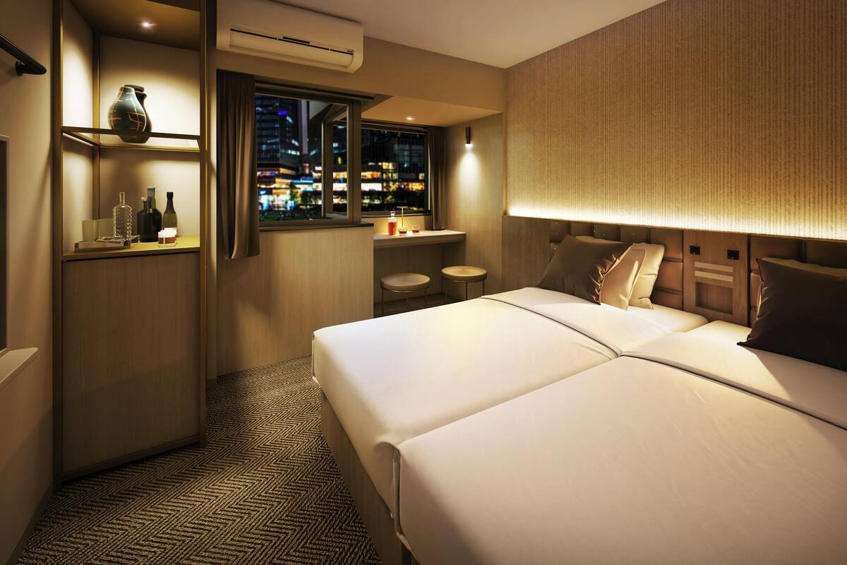 A hotel room with a bed, a desk with a vase, bottles, and a city view through the window.
