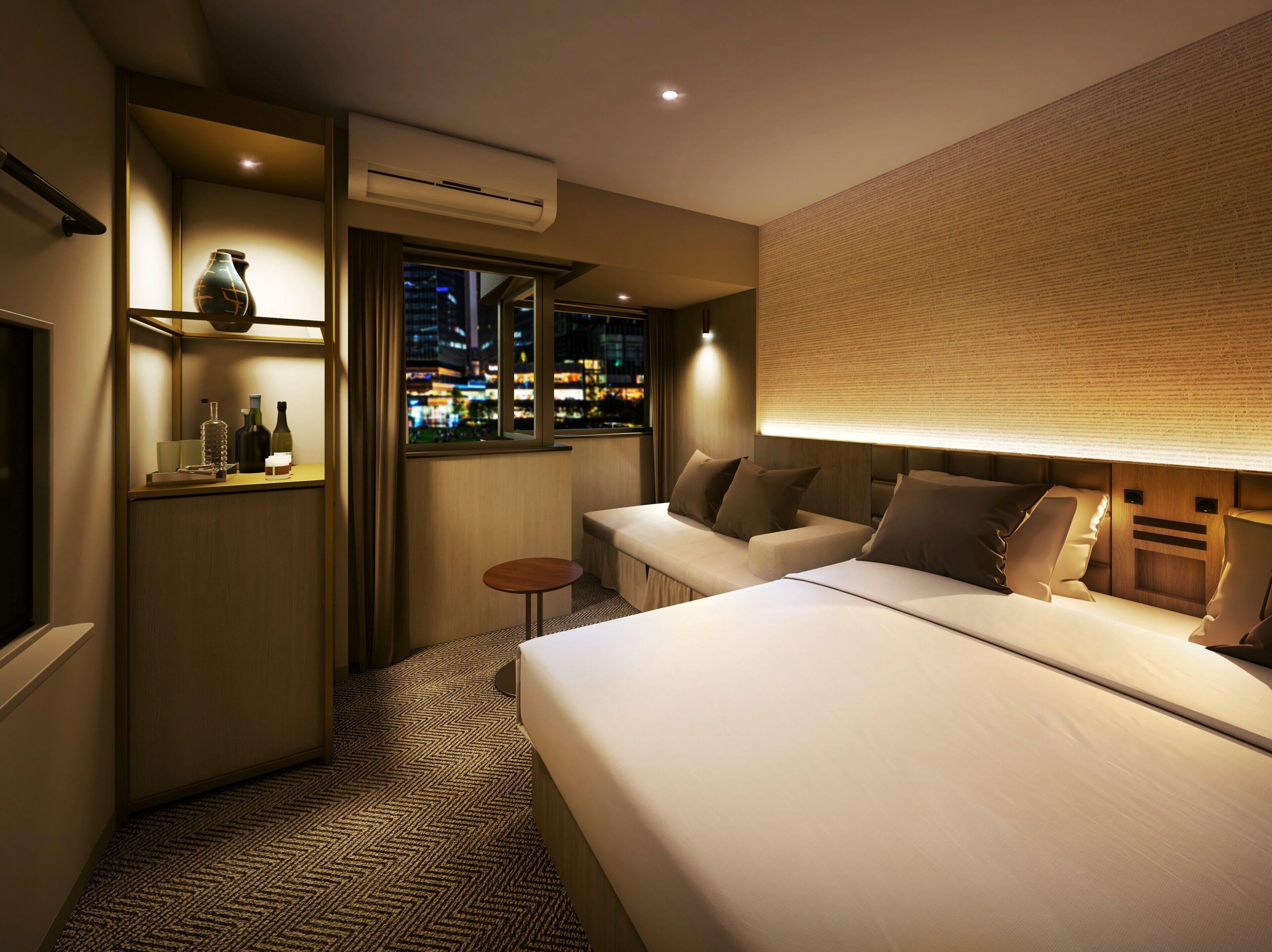 A hotel room with a bed, a seating area, a small table, an air conditioning unit, and a view of the city at night.