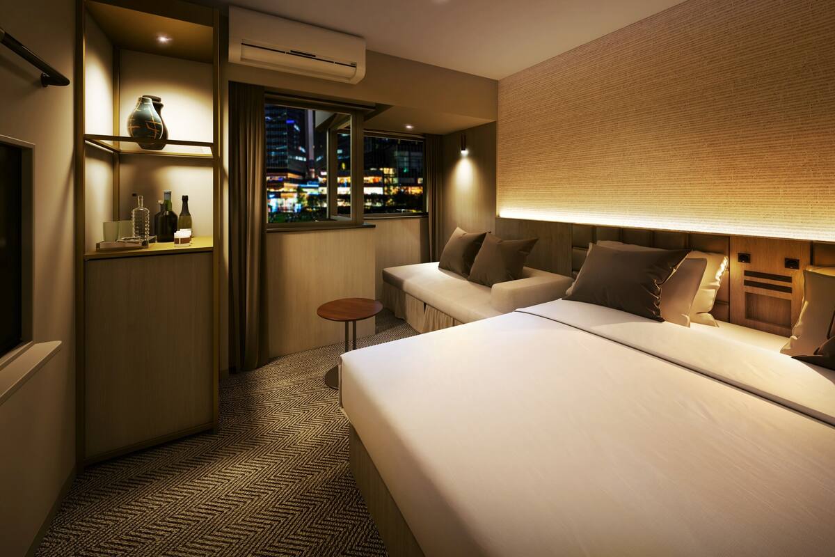 A hotel room with a bed, a seating area, a small table, an air conditioning unit, and a view of the city at night.