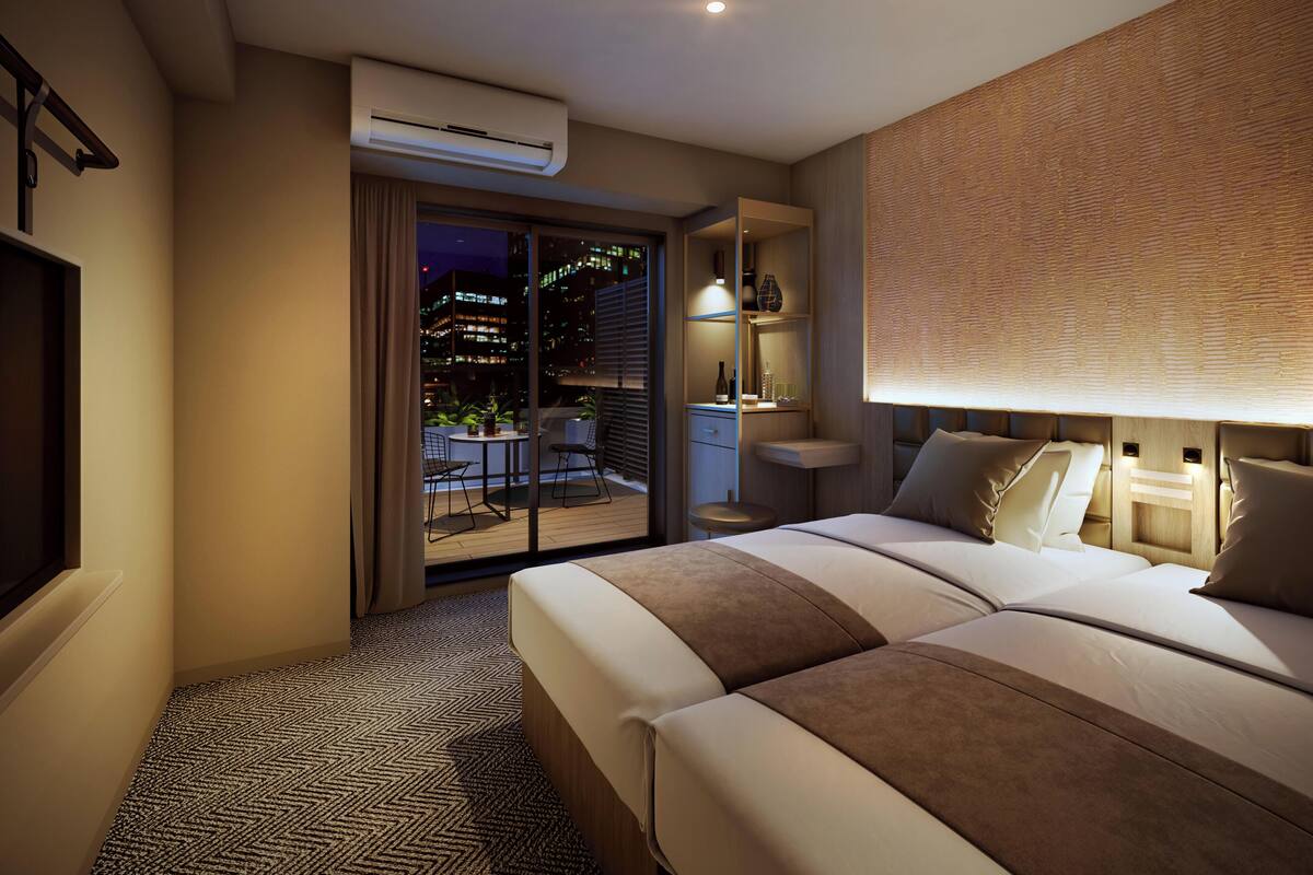 A hotel room with a bed, a desk, a chair, an air conditioning unit, and a view of the city at night.