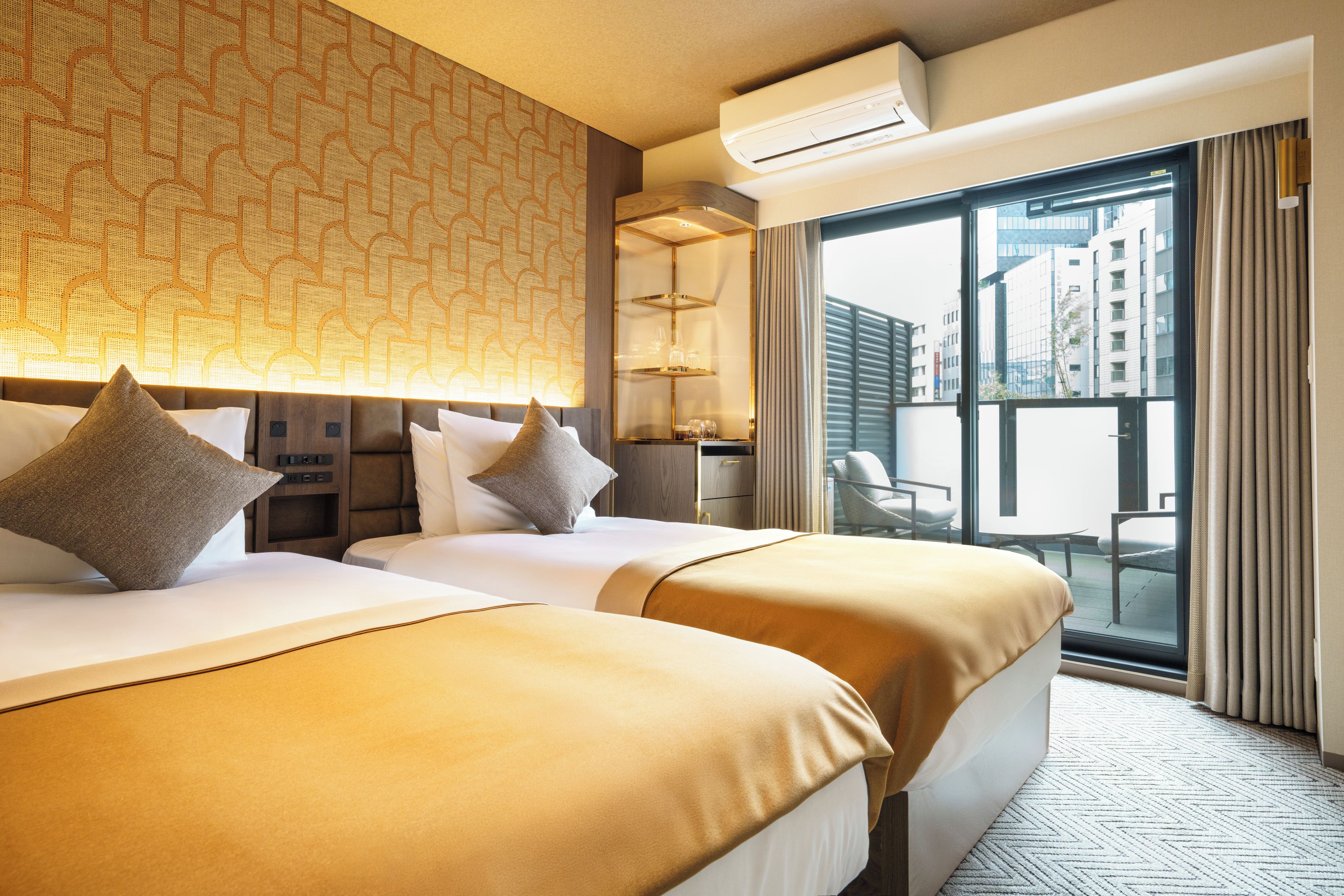 A hotel room with two beds, a minibar, a large window with a city view, and an air conditioning unit.