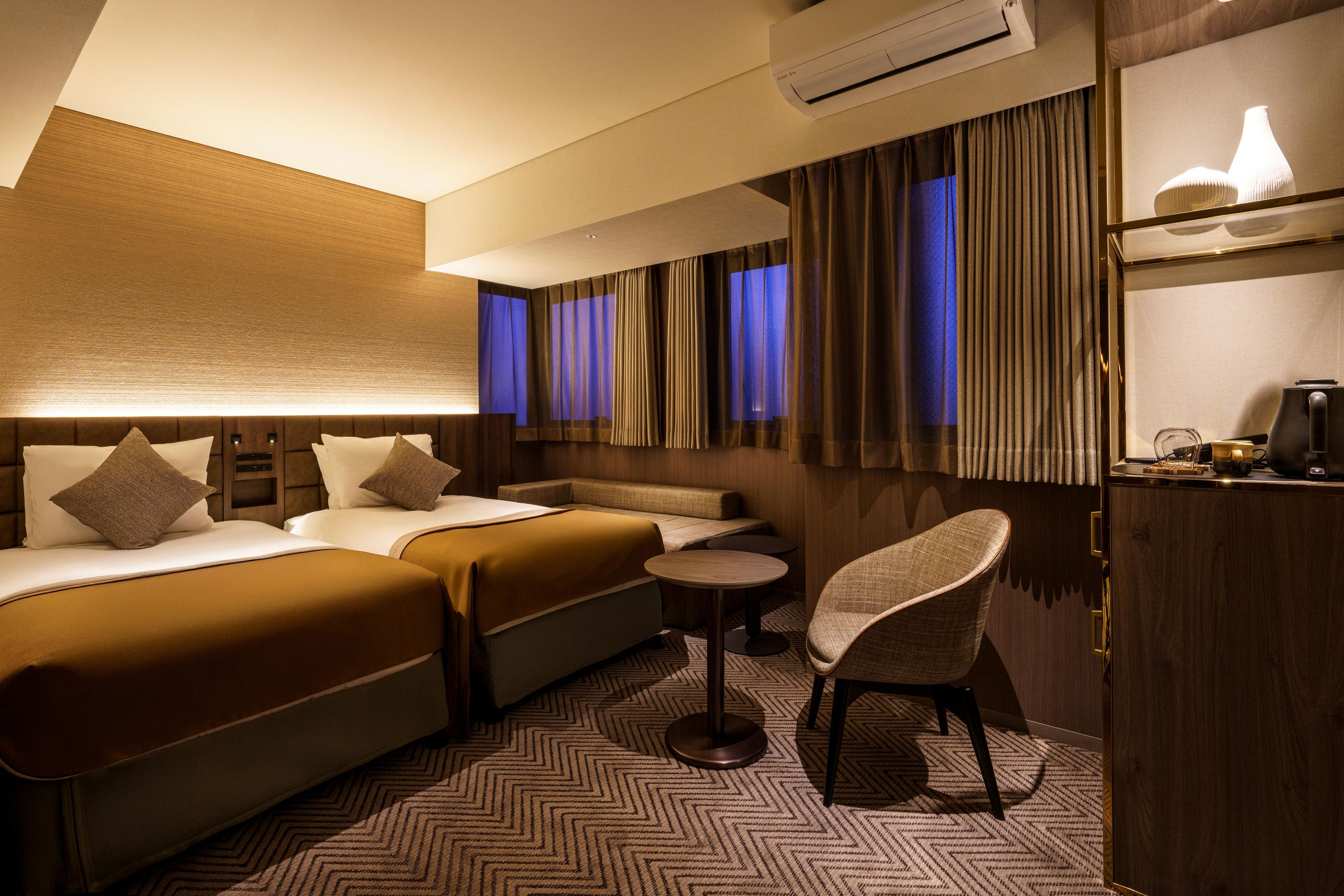 A hotel room with a bed, a sofa, a chair, an air conditioning unit, and a small table.