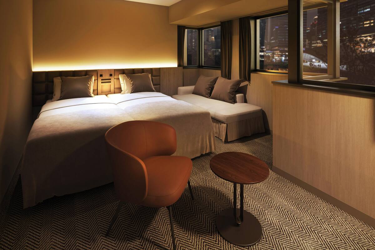 A hotel room with a large bed, a sofa, a chair, and a small table. There are multiple windows providing a view of the city at night.