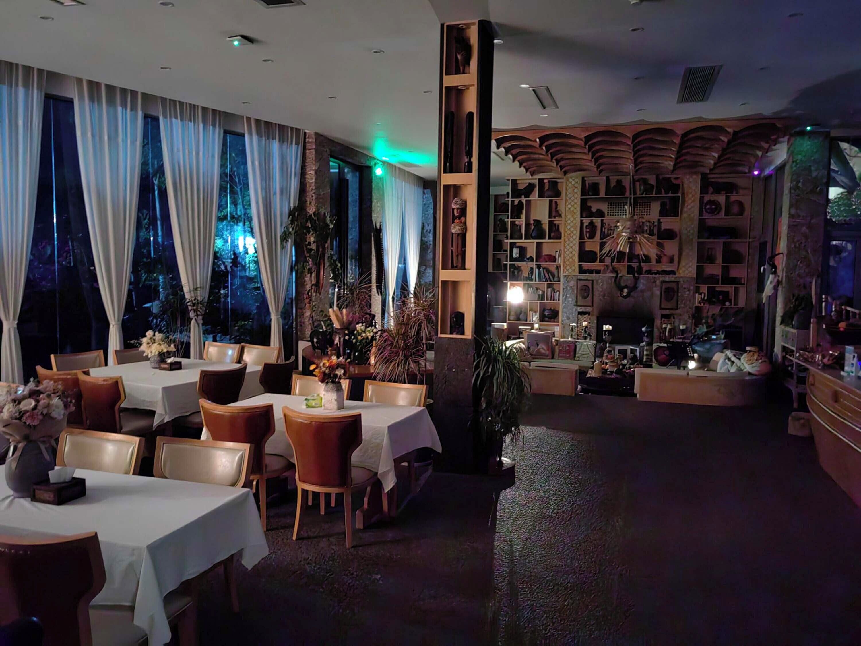 Un restaurante con mesas y sillas, una estantería y un ventanal con cortinas.