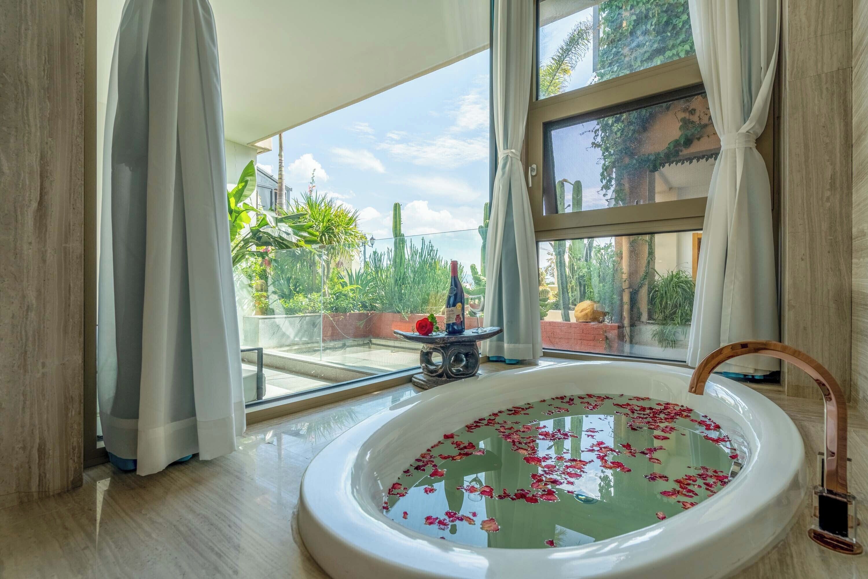 Une salle de bain avec une baignoire remplie d’eau et de pétales de rose, une fenêtre avec des rideaux et une vue sur la verdure à l’extérieur.