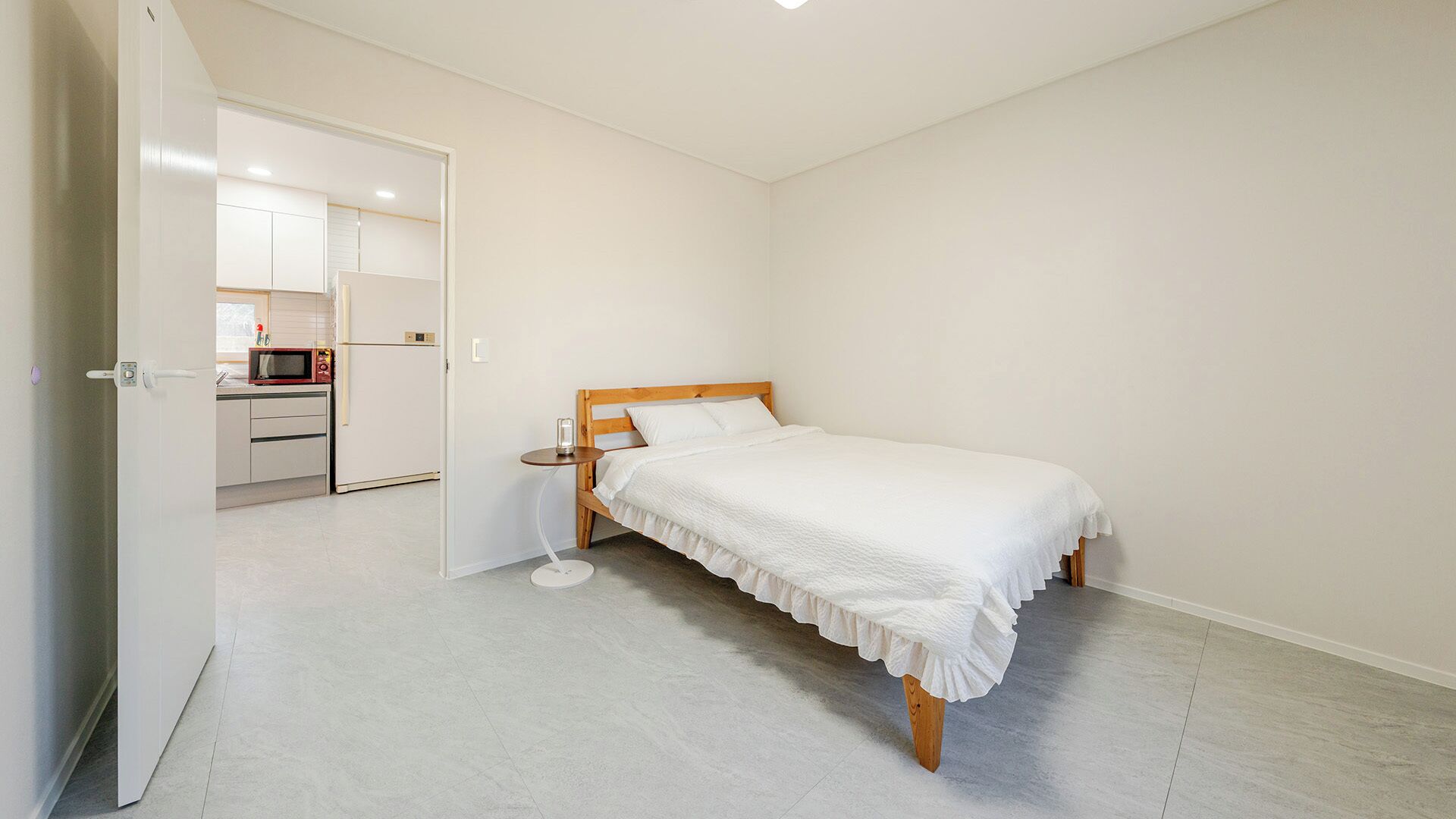 Ein Schlafzimmer mit einem Einzelbett, einem kleinen Nachttisch und einem Beistelltisch. Eine Tür führt zu einer Küchenzeile mit einer Mikrowelle und einem Kühlschrank.