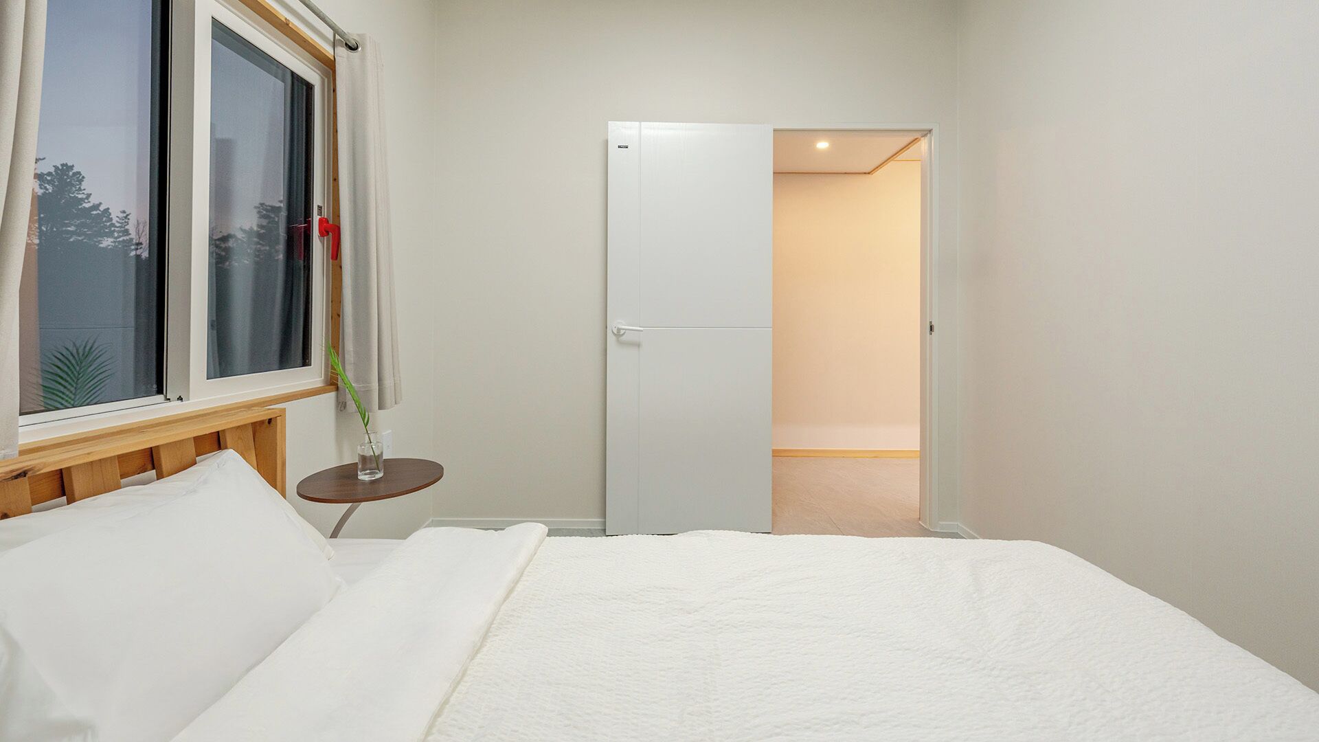 Une chambre à coucher avec un lit, une fenêtre avec des rideaux, une table de chevet avec une plante, et une porte donnant sur une autre pièce.