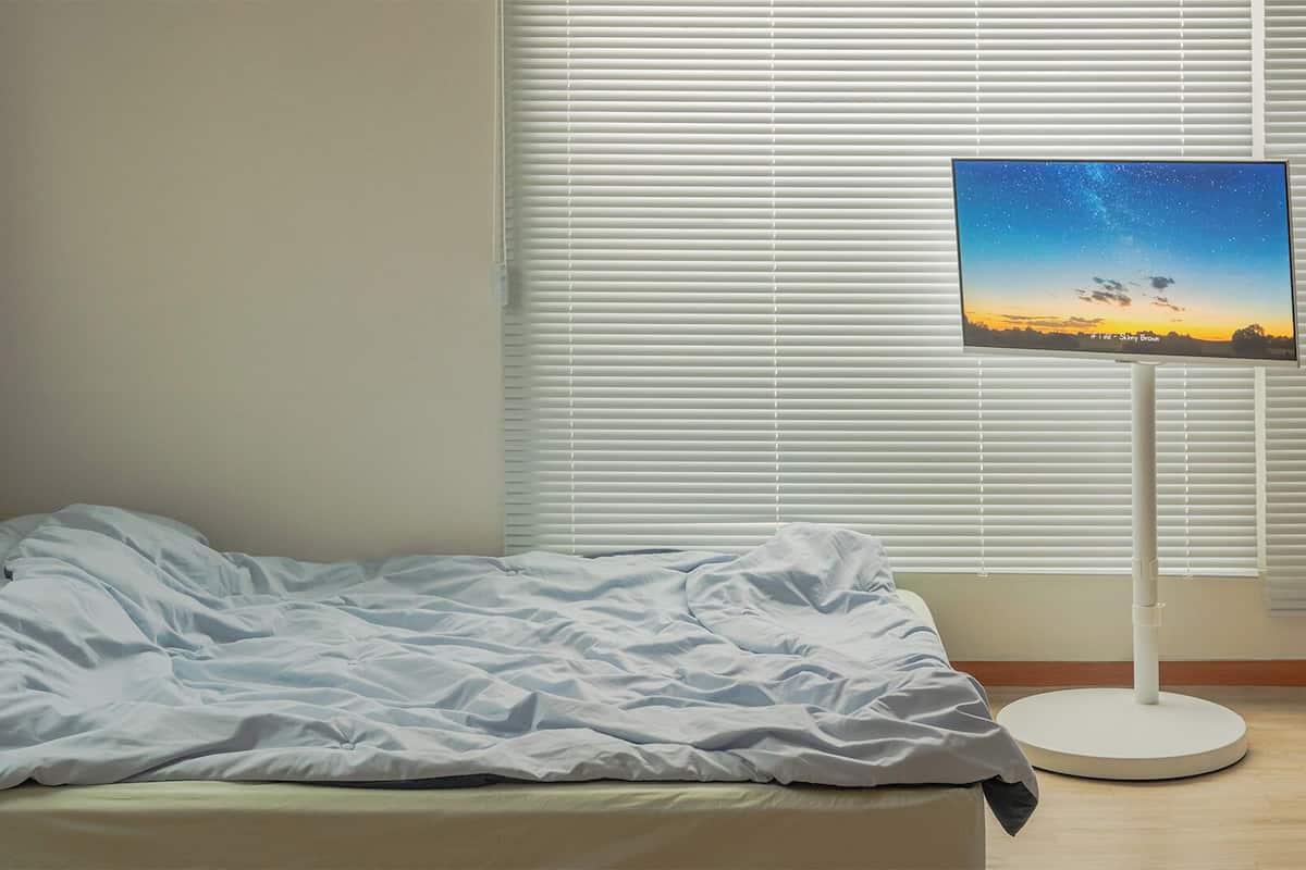 Una cama con edredón blanco, una mesita de noche y un televisor sobre un soporte que muestra un cielo nocturno.