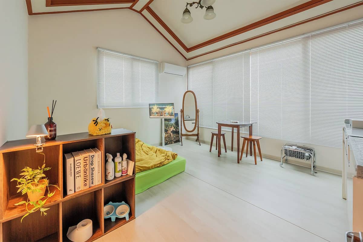 Una habitación con una estantería, un sofá verde, una mesa con sillas y ventanas grandes con persianas.