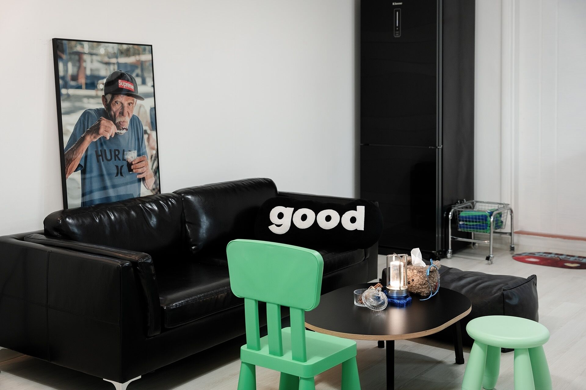 Uma sala de estar moderna com um sofá de couro preto, uma poltrona verde, uma mesa redonda pequena com objetos decorativos e um quadro na parede.