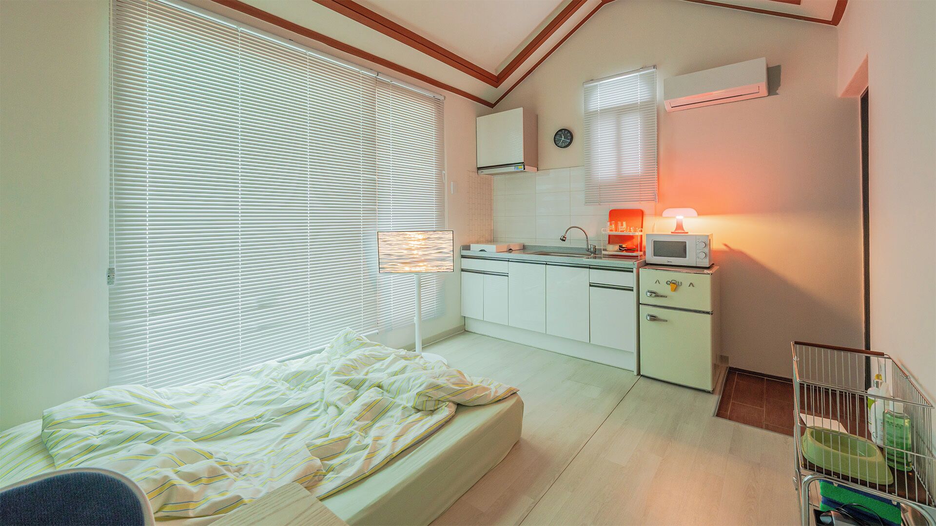 Une chambre comprenant un lit, une table de chevet, une fenêtre avec des stores, une kitchenette équipée d’un four à micro-ondes et un petit placard.