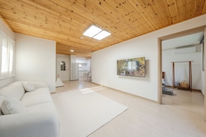 Un salón amplio con techo de madera, un sofá blanco, un cuadro grande y vistas hacia una cocina.