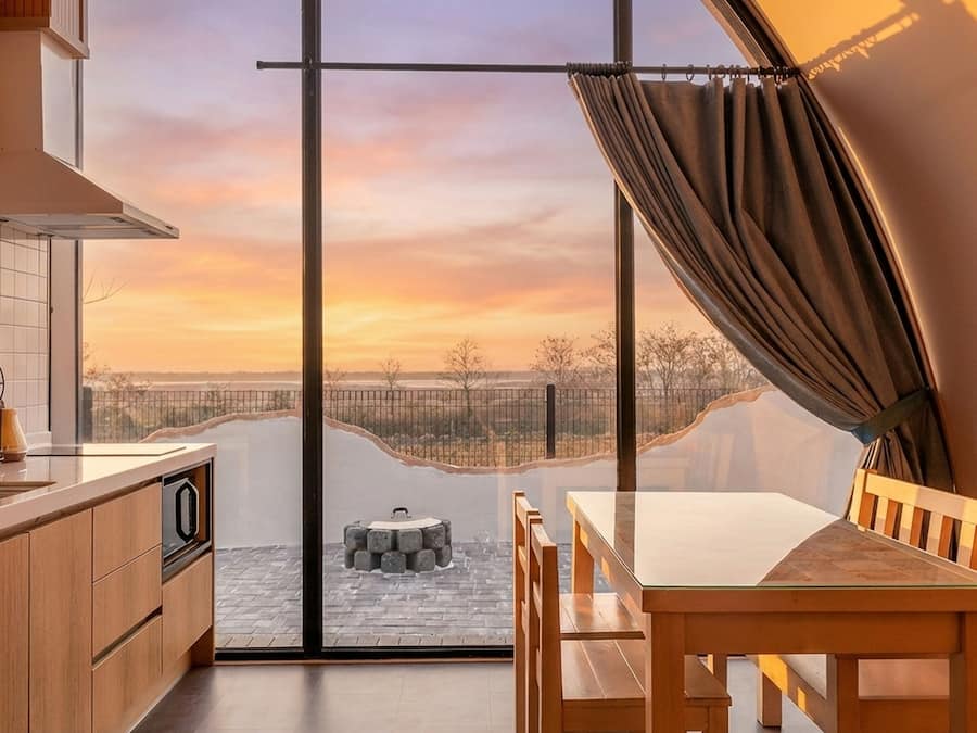 A modern kitchen with a large sliding glass door leading to a balcony with a view of a sunset over a distant landscape.