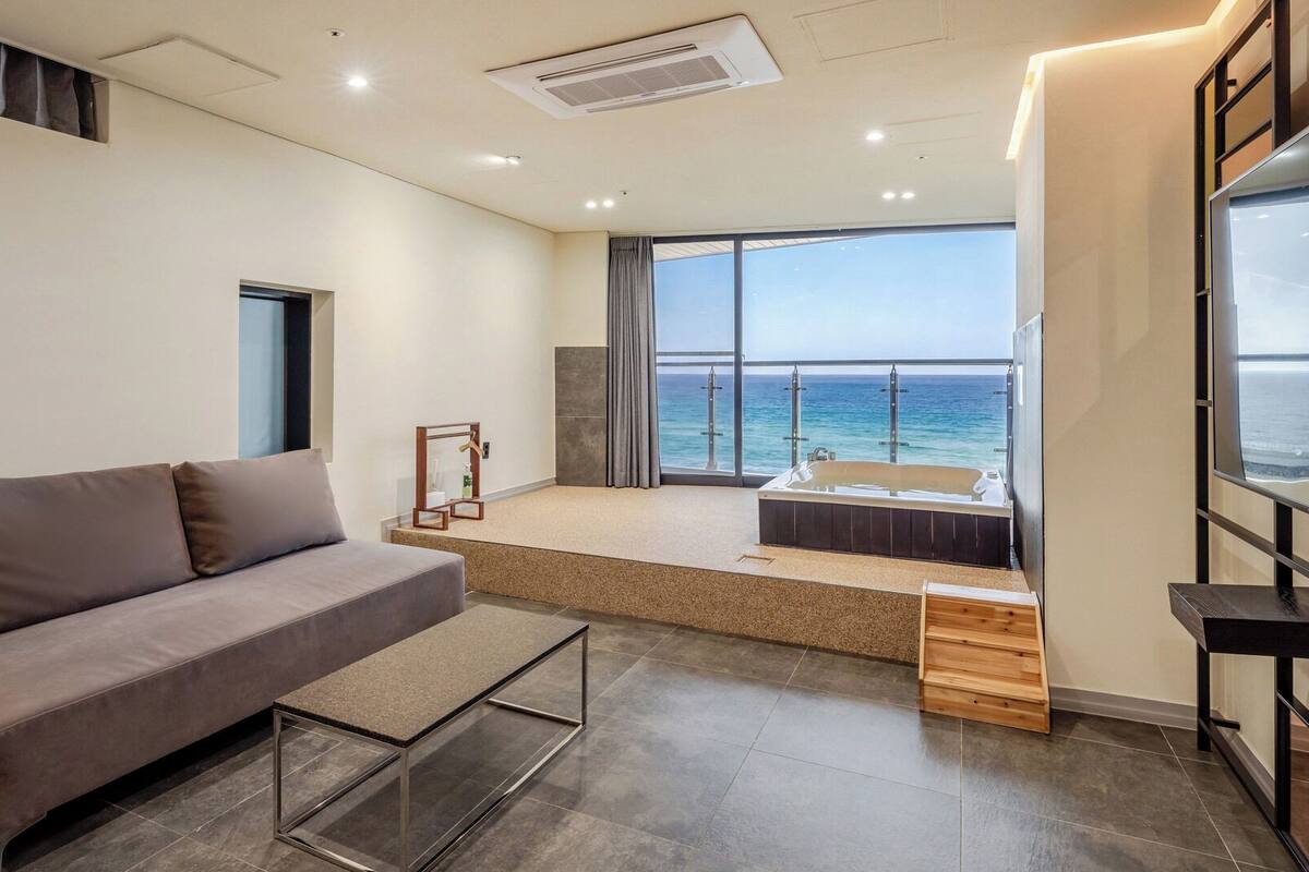 A modern living room with a sofa, a small table, a view of the ocean, and a balcony with a bathtub.