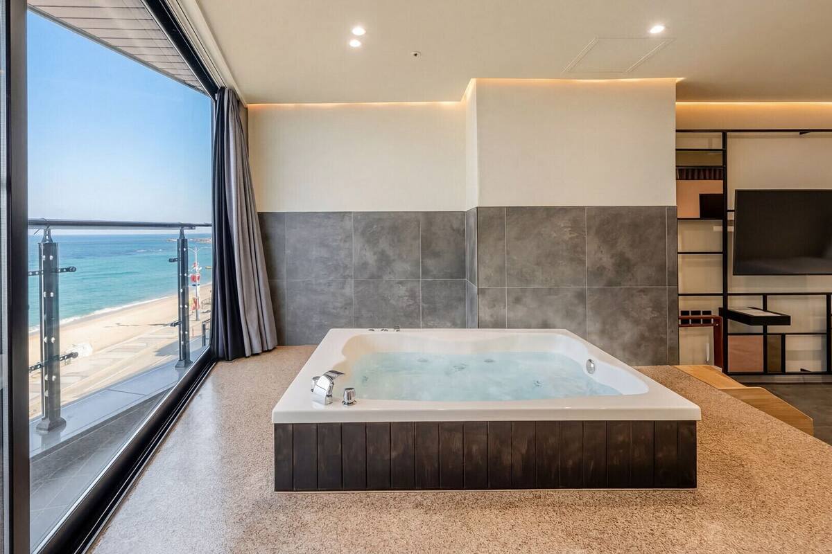 A modern bathroom with a large bathtub, a glass shower enclosure, and a view of the beach.