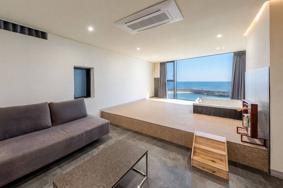 A modern hotel room with a sofa, a small window, a bathtub, and a view of the sea.