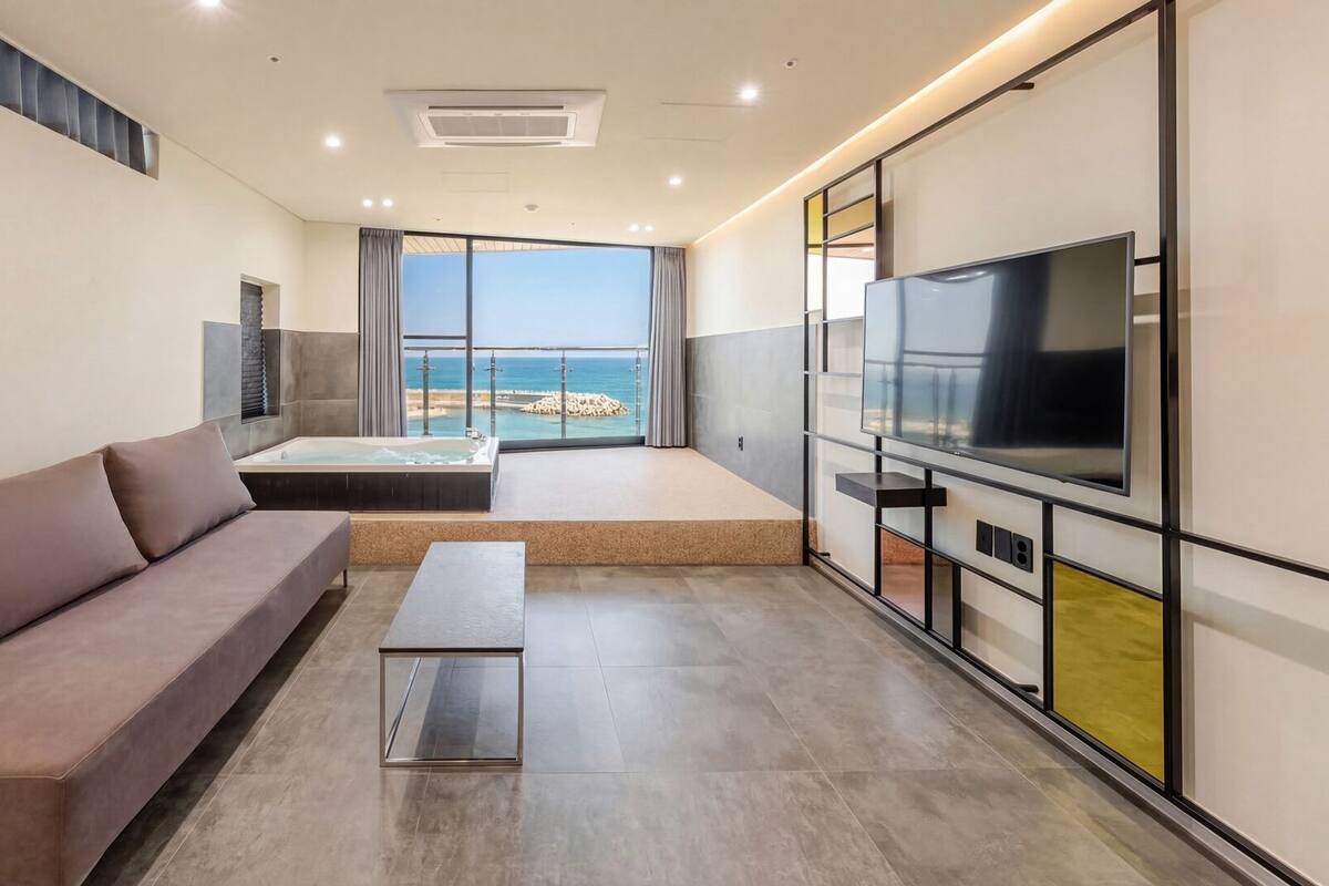 A modern hotel room with a sofa, a small table, a TV, and a balcony with a view of the sea.