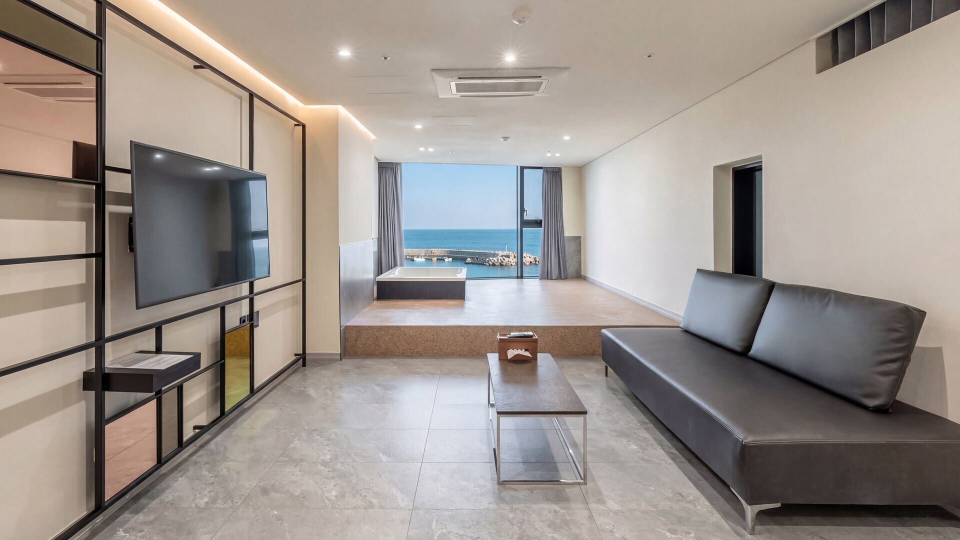 A modern living room with a flat-screen TV, a sofa, a coffee table, and a view of the sea.