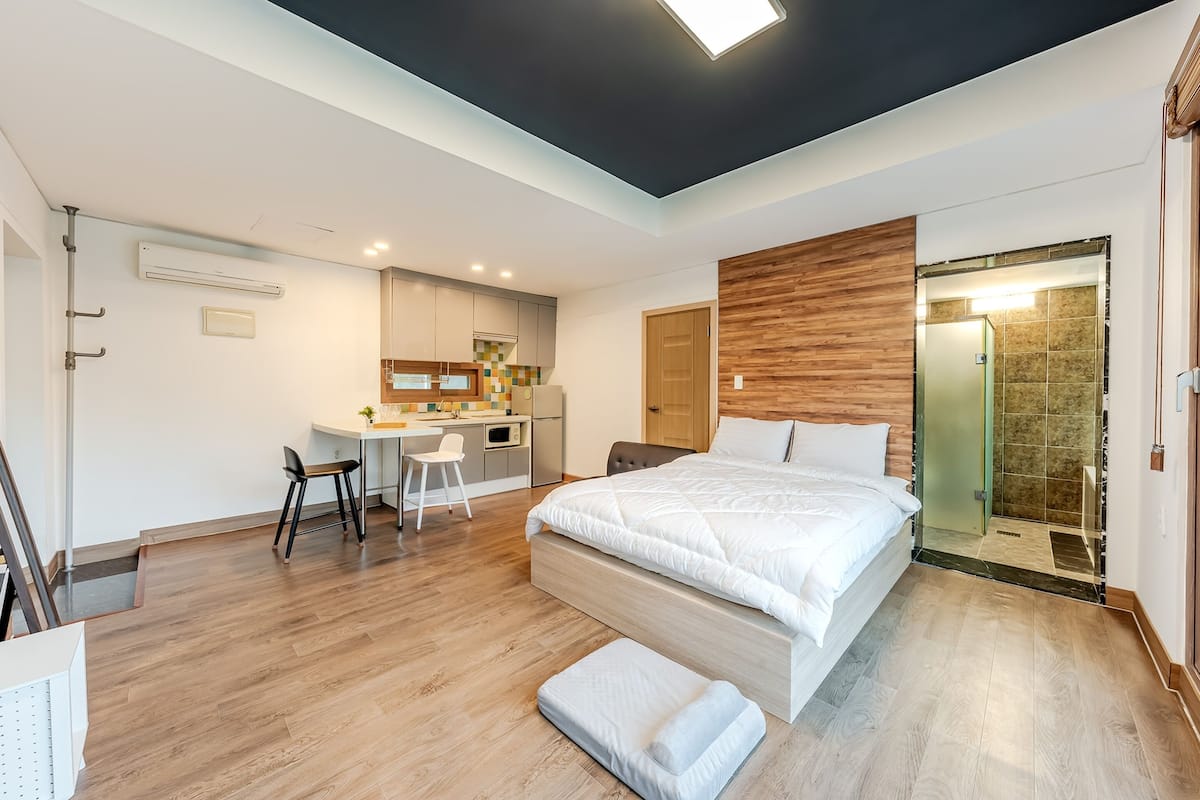Ein modernes Hotelzimmer mit einem großen Bett, einem Schreibtisch mit Stuhl, einer kleinen Küchenzeile und einem Duschbereich.